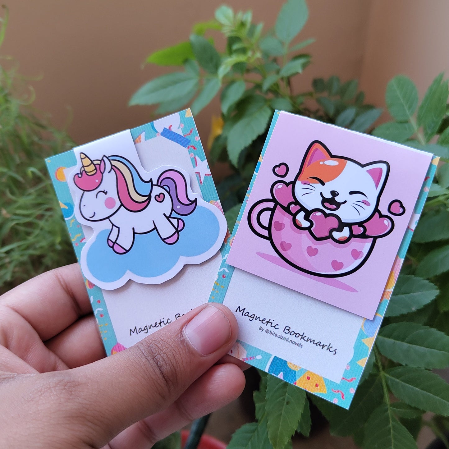 A hand holding a customizable magnetic bookmark with a colorful design featuring an illustration of a cat on a unicorn-themed background.